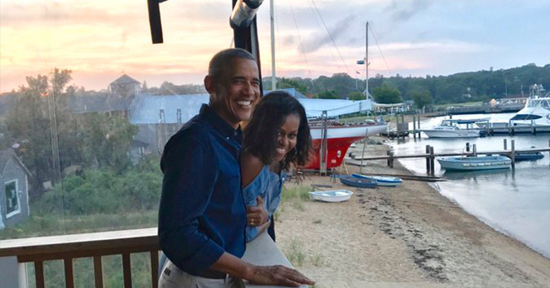 Barack and Michelle Obama laughing on a porch near some boats and water
