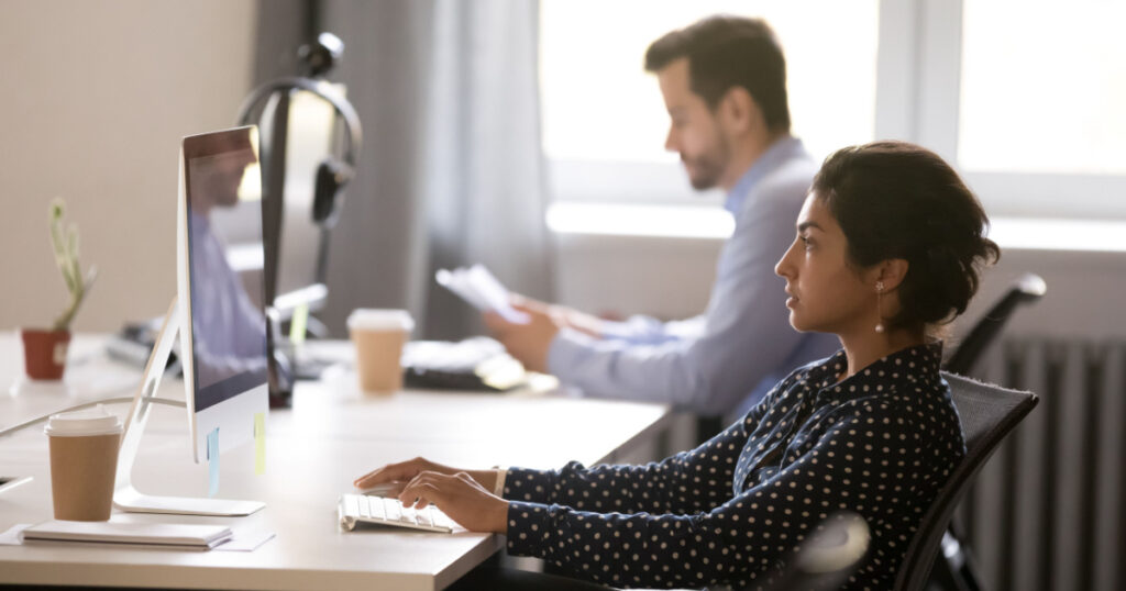 Side view diverse employees sitting at desk using laptop, focus on young indian female. Millennial serious hindu worker typing message business letter on computer looking at screen searching new ideas