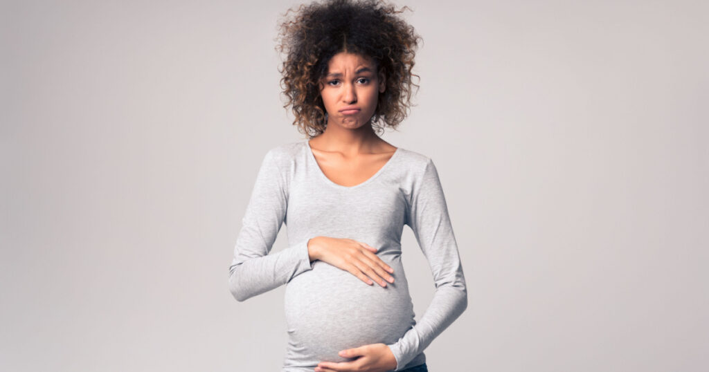 Hormones and mood changes. Sad expectant woman touching belly over grey studio background