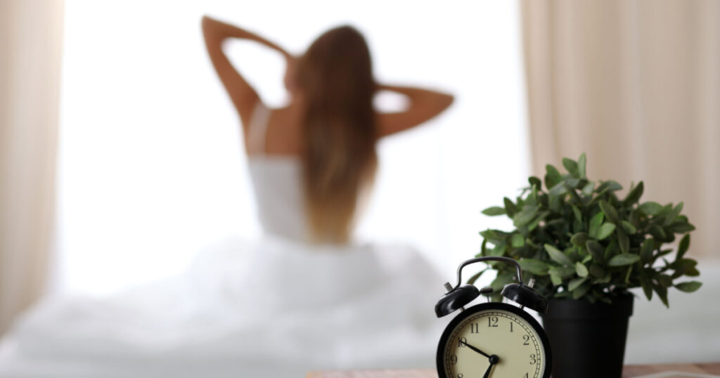 Alarm clock standing on bedside table. early morning wakes up woman, stretching in bed in background