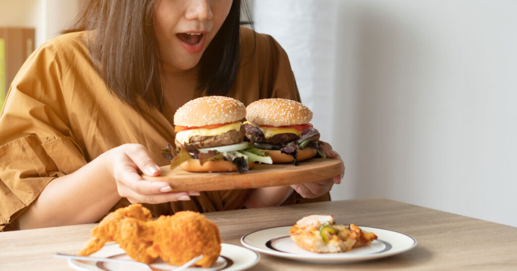 Hungry overweight woman holding hamburger on wooden plate, Fried chicken and Pizza on table