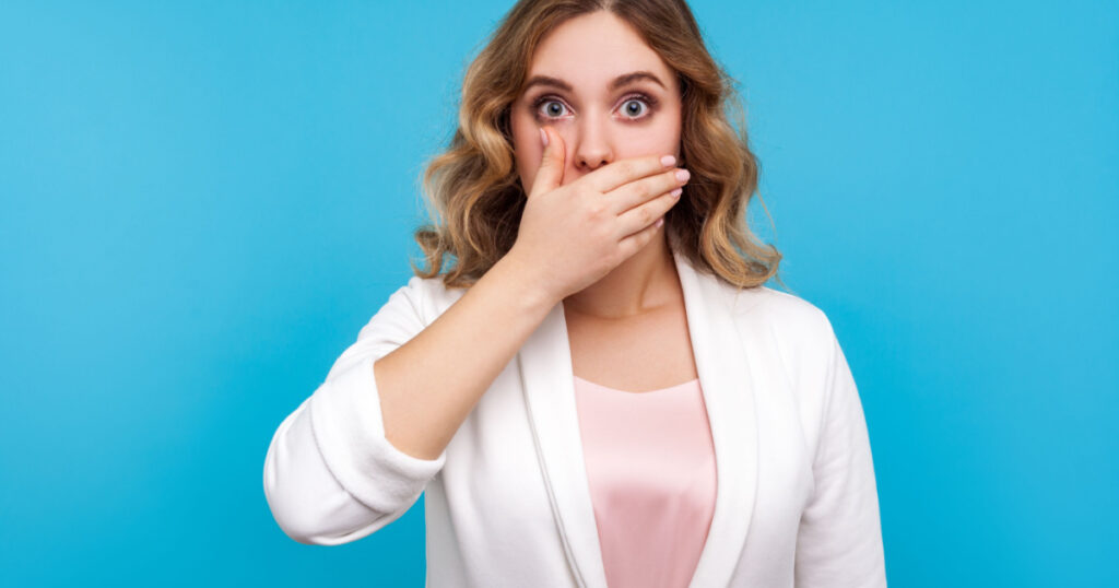 Portrait of frightened woman with wavy hair in white jacket covering mouth with hand scared