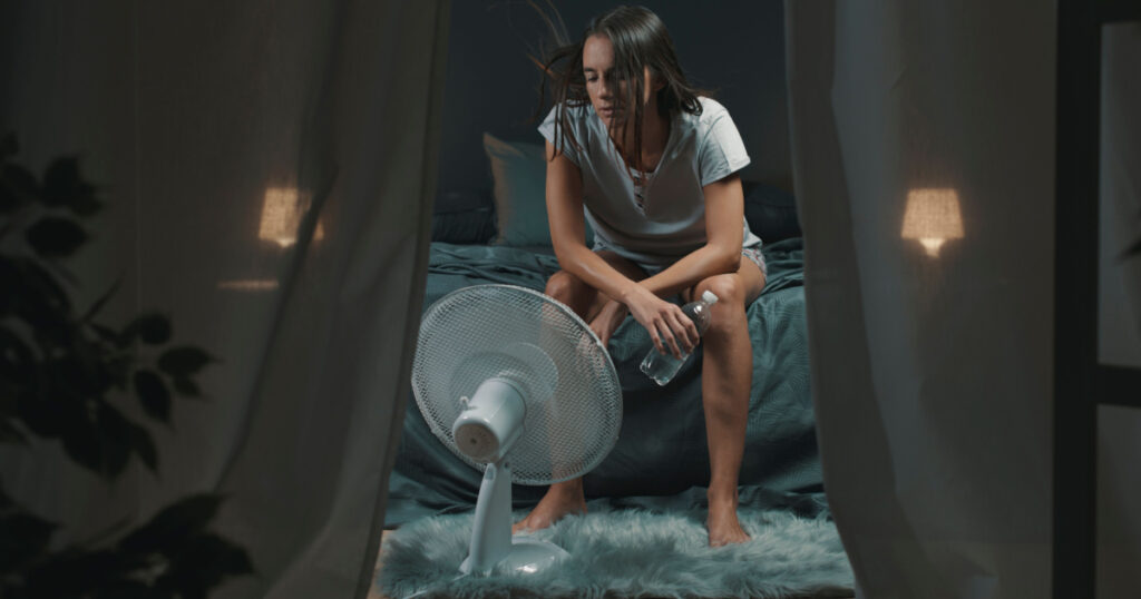 Exhausted woman suffering suring the heatwave, she is holding a water bottle and sitting in front of a cooling fan in the bedroom