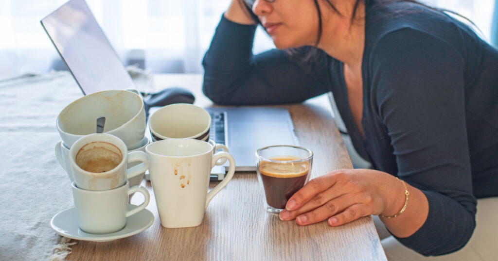 Asian woman holding a cup of coffee sitting tired with many empty cups of coffee and laptop on the desk