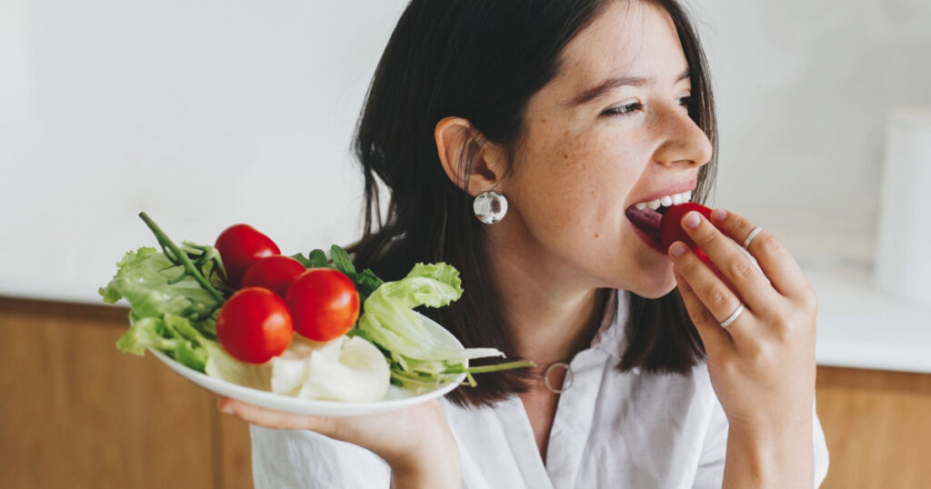 woman eating cherry tomato and holding plate with fresh green lettuce, cherry tomatoes, arugula in modern kitchen