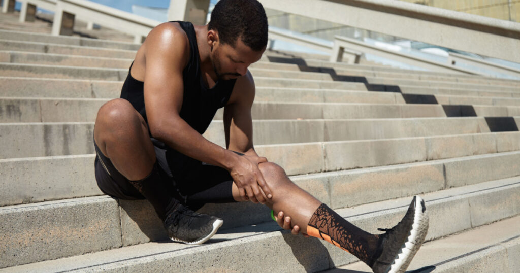 runner with muscular athletic body holding his leg with both hands, feeling pain in knee or calf, massaging it, suffering from strain or spasm while sitting on steps of concrete stair