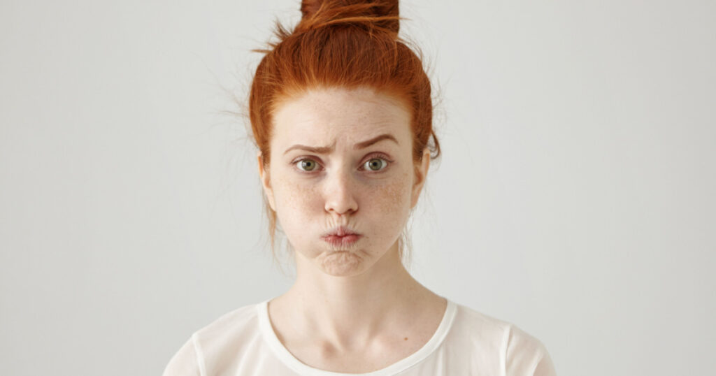Annoyed irritated young red-haired female with freckles blowing her cheeks, frowning, feeling frustrated 