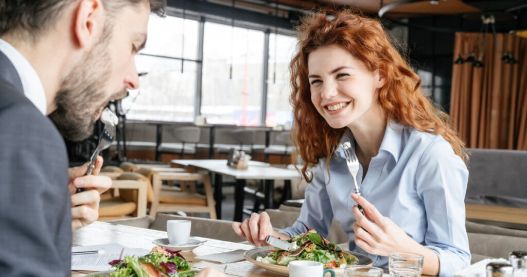 Man and woman having business lunch at restaurant sitting at table businessman eating vegetable salad with feta cheese and salmon concentrated businesswoman laughing joyful
