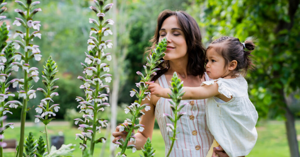 Side view of relaxed mother with active child in hands smelling flowers with closed eyes and enjoying fragrance in green park
