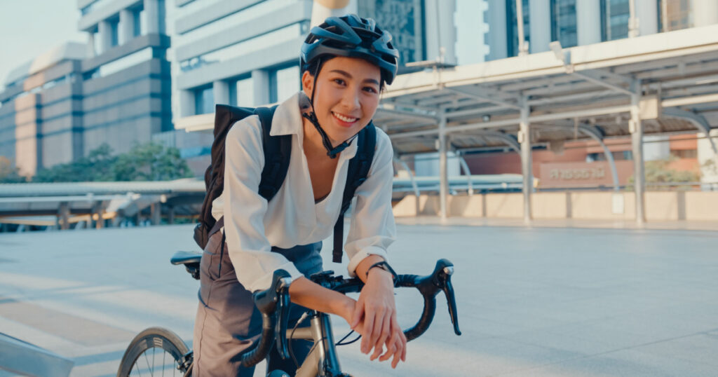 Asian businesswoman go to work at office stand and smiling wear backpack look at camera with bicycle on street around building on a city. Bike commuting, Commute on bike, Business commuter concept.
