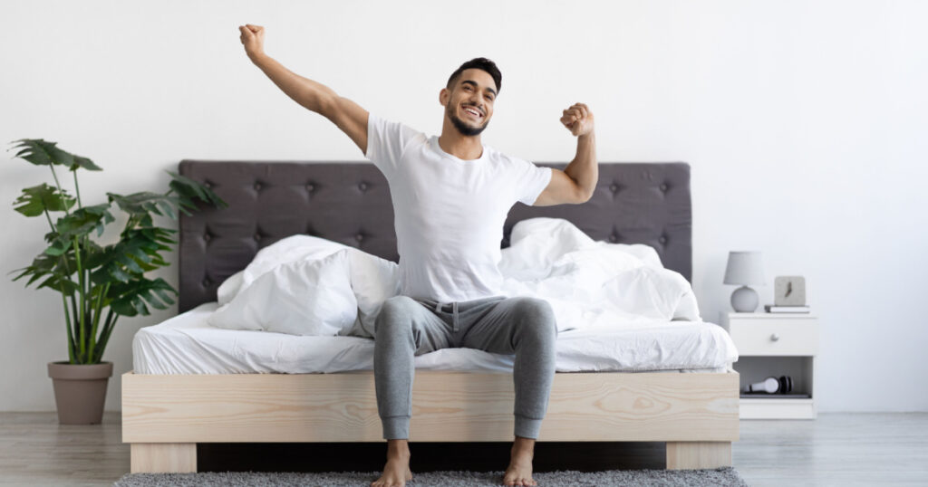 Guy Waking Up In The Morning, Sitting On Bed And Stretching After Good Sleep,