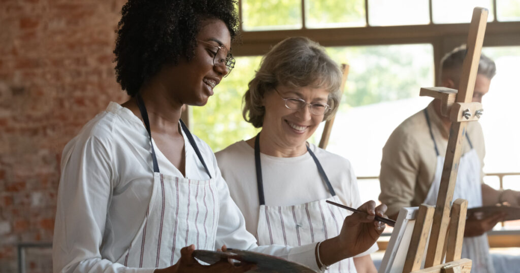 Happy young African American woman in eyeglasses taking advices from skilled old senior female teacher or friend, discussing artwork drawing with brushes on canvas, enjoying creative art class.
