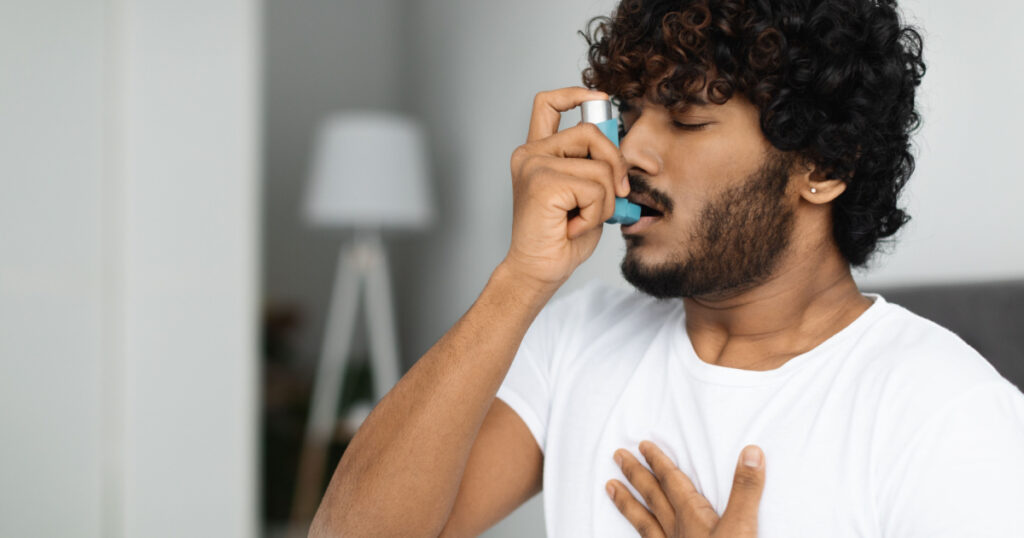 Indian guy suffering from asthma, using inhaler in bed, side view, copy space. Young hindu man woke up with asthma attack, touching chest and using nebulizer, bedroom interior
