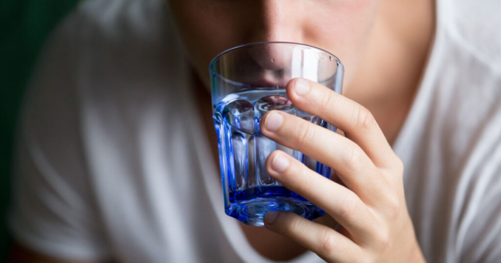 Close up of young man feeling thirst holding transparent blue glass drinking still water, focus on arm. Dehydration of organism or health care, good habit and weight loss. Hangover after party concept
