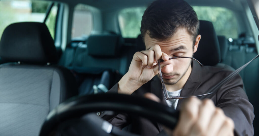 Sleepy young man rubs his eyes with his right hand. His left hand is on the steering wheel. He is sitting at his car. Road safety concept.
