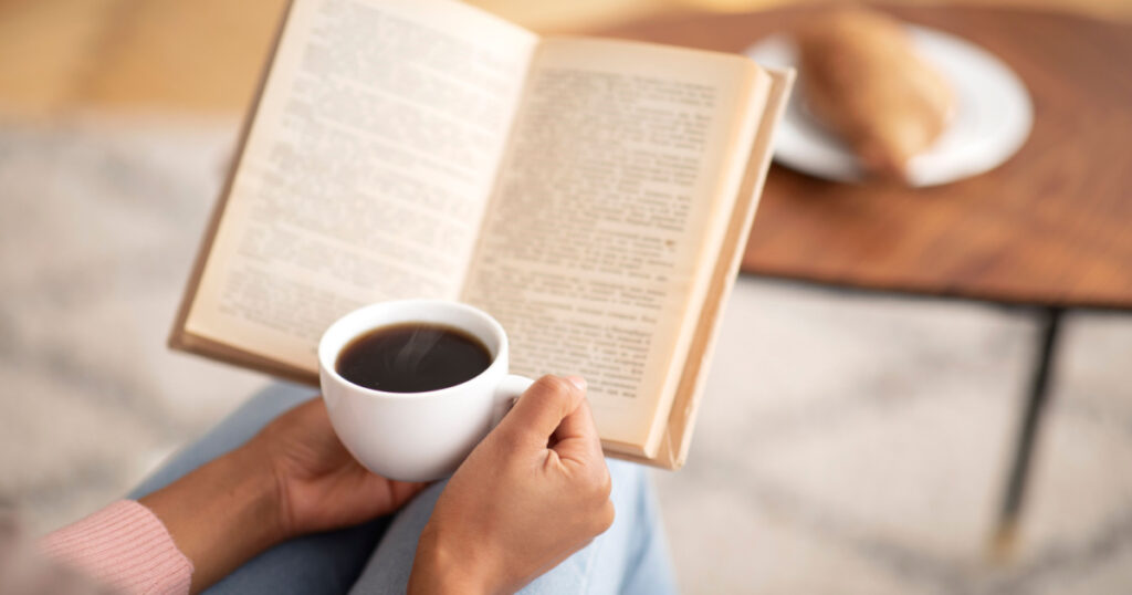 Unrecognizable black lady with coffee reading book, enjoying peaceful morning at home, free space. Cropped view of female hands holding hot beverage and textbook on weekend, indoors
