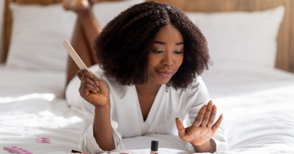 Pretty black woman lying on bed with nail file, doing manicure, taking care of her hands at home. Charming African American lady making self-care procedure indoors. Wellness routines concept
