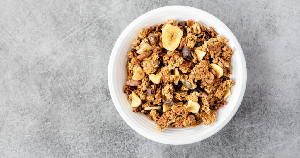 Homemade granola in a white bowl on a white background, overhead view. Homemade whole grain musli with bananas and dark chocolate for breakfast. Top view, light, airy, clean. Breakfast cereal.
