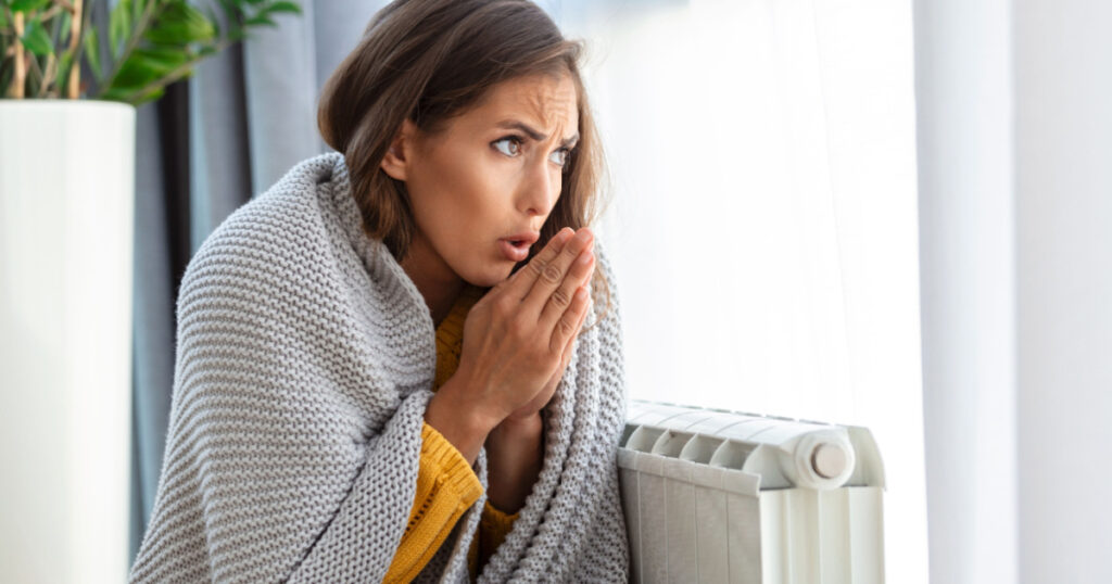 Unwell woman renter in blanket sit in cold living room hand on old radiator.suffer from lack of heat . Unhealthy young woman struggle from chill freeze at home. No heating concept.
