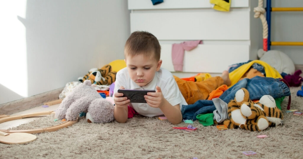 boy (5-6 years old) in bedroom, uses phone, plays phone games in dirty children's room, child plays among lot of toys at home,lot of toys and things scattered on floor of the room, a dirty house.
