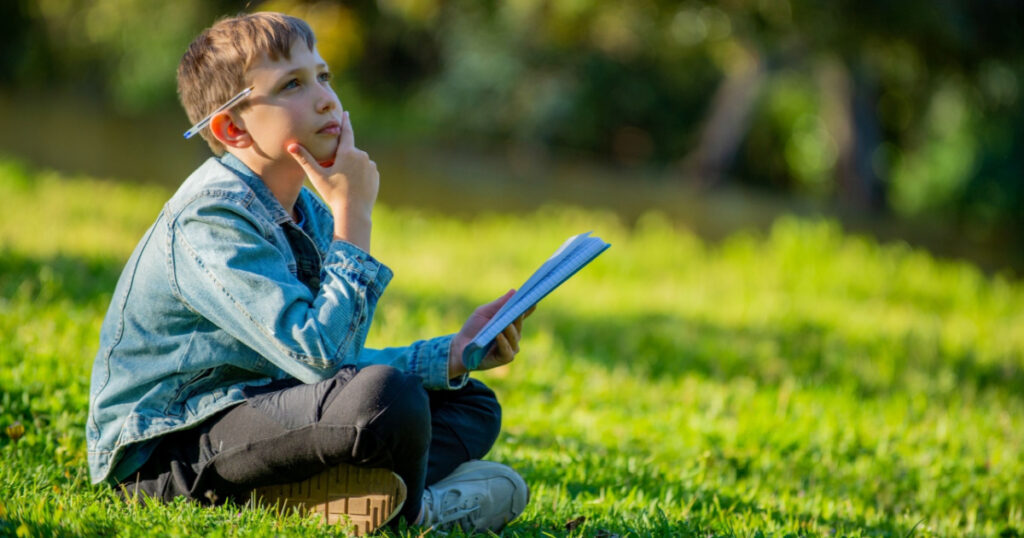 Thinking student with a textbook in his hands is sitting on a green lawn in a park. Young boy reading his notes and memorizing the text before a test. A student writing in a notebook in a garden
