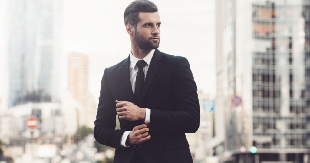 Modern businessman. Confident young man in full suit adjusting his sleeve and looking away while standing outdoors with cityscape in the background
