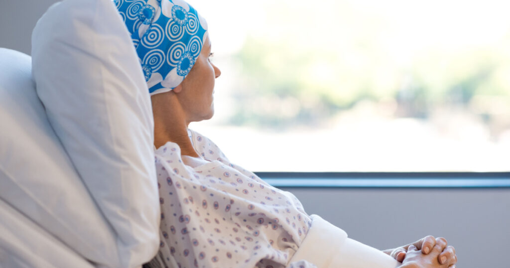 Young woman in bed suffering from cancer. Thoughtful woman battling with tumor looking out of window. Young patient with blue headscarf recovery in hospital on bed.
