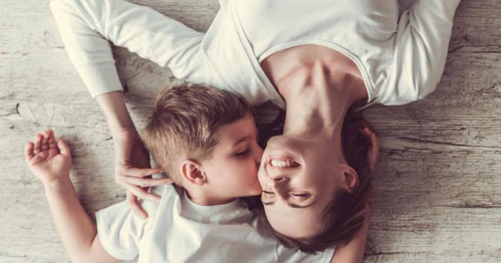 Top view of beautiful woman and her cute little son smiling while lying on the floor. Boy is kissing his mom in cheek
