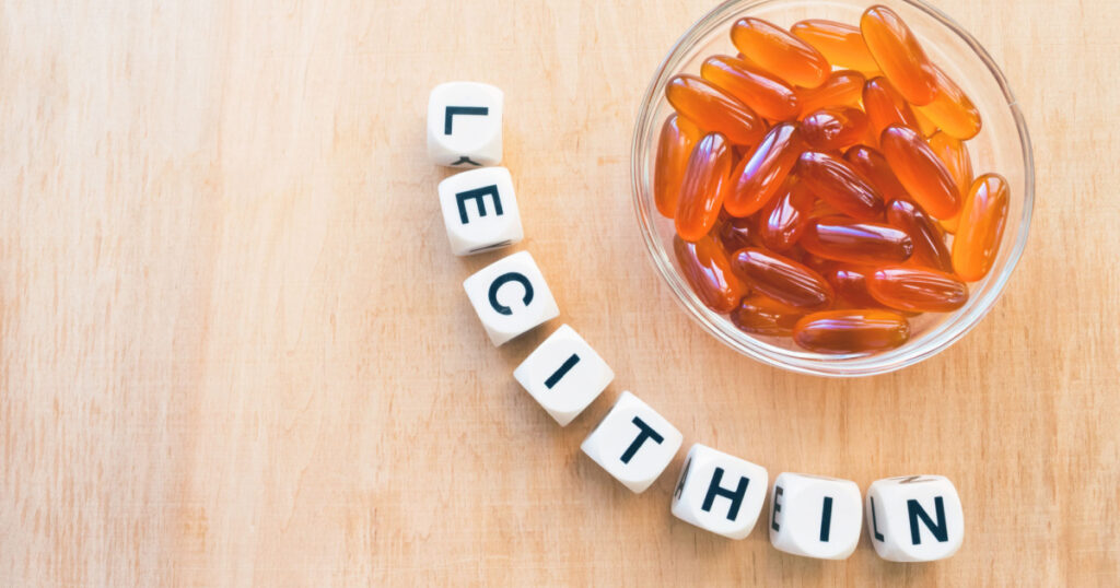 Lecithin gel pills in a round glass bowl and with the word Lecetin from the letters of cubes on a light wooden background. Soy and sunflower lecithin benefits for skin, digestion, lower cholesterol.
