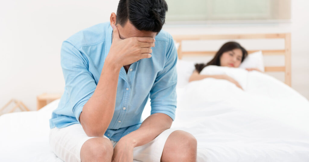 Husband unhappy and disappointed in the erectile dysfunction during sex while his wife sleeping on the bed. Sexual Problems in Men.
