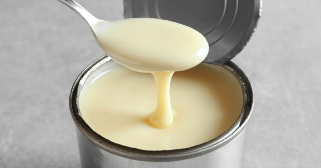 Condensed milk pouring from spoon into tin can on grey background, closeup. Dairy product
