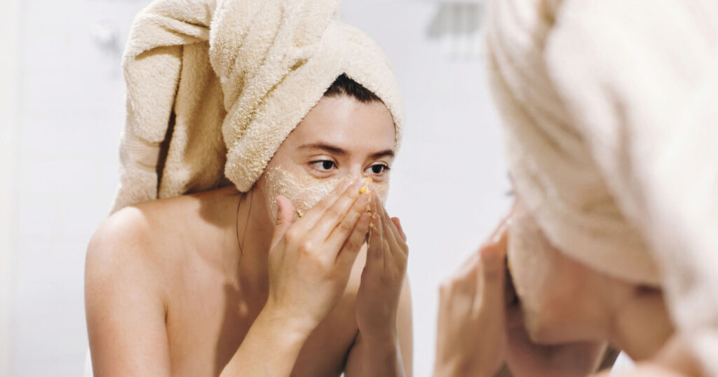 woman in towel making facial massage with organic face scrub and looking at mirror in stylish bathroom