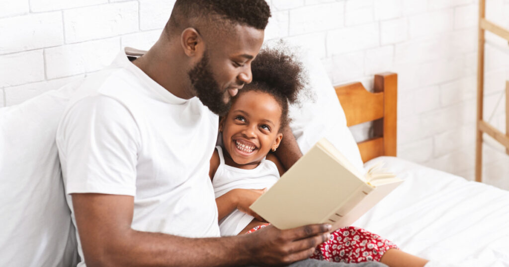 Happy evening. African american man reading fairy tale to his little daughter, sitting in bedroom, empty space
