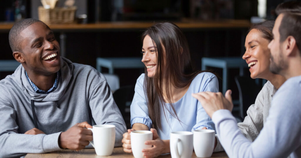 Multiracial friends girls and guys having fun laughing drinking coffee tea in coffeehouse, happy diverse young people talking joking sitting together at cafe table, multicultural friendship concept
