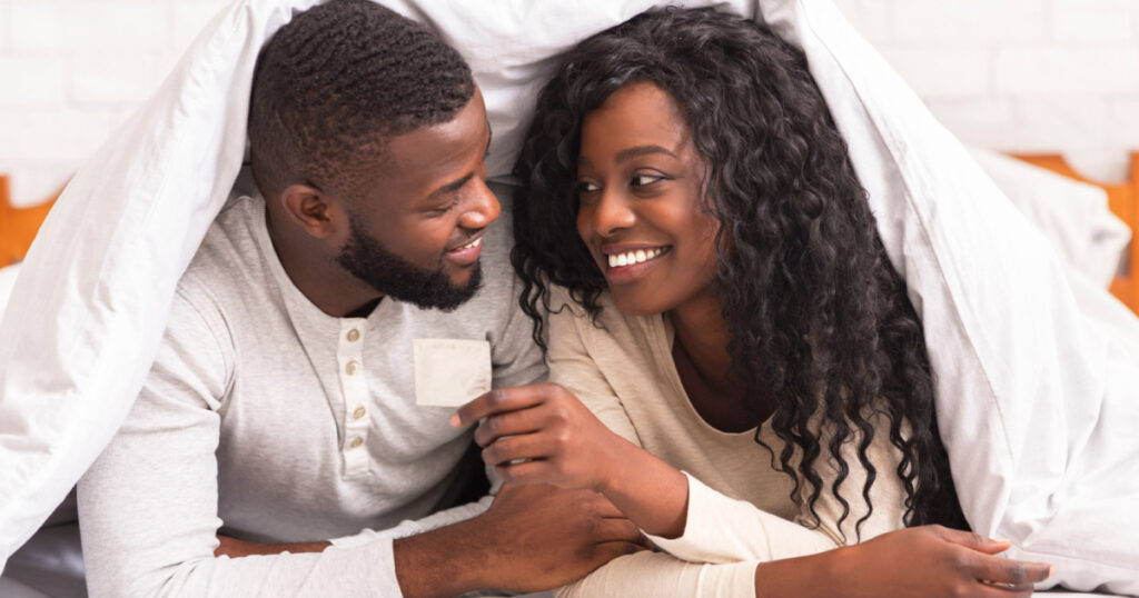 Safe sex. Young african american couple holding condom, lying on bed under white blanket, looking at each other with love and care
