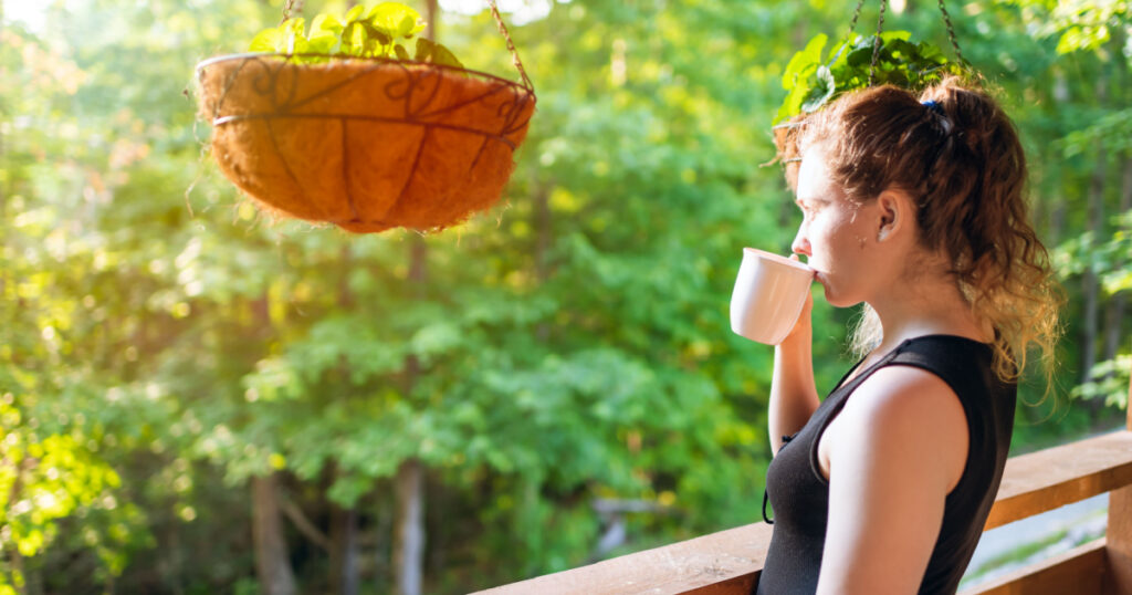 Hanging potted plants in summer with woman standing on porch of house in morning wooden cabin cottage drinking tea or coffee
