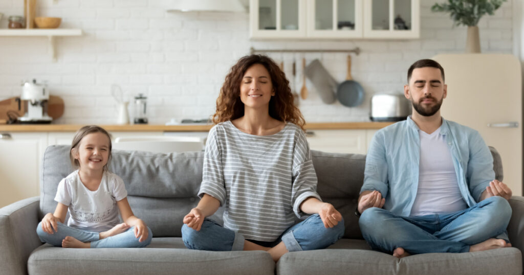 Calm young family with little daughter sit on couch practice yoga together, happy parents with small preschooler girl child rest on sofa meditate relieve negative emotions on weekend at home
