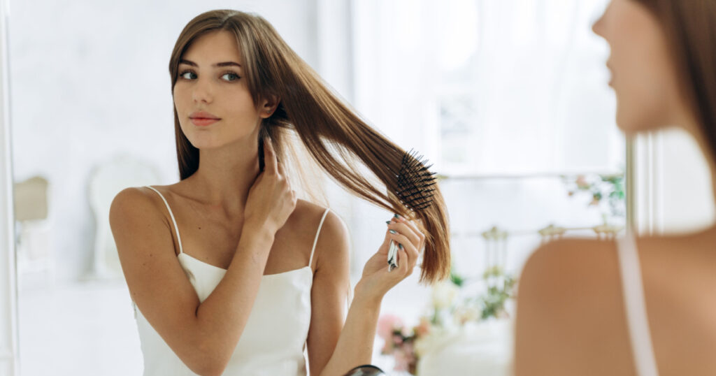Waist up portrait of the woman reflected in mirror doing daily routine while holding hairbrush tidy her hair. Female looking and tangled hair. Beauty treatment concept
