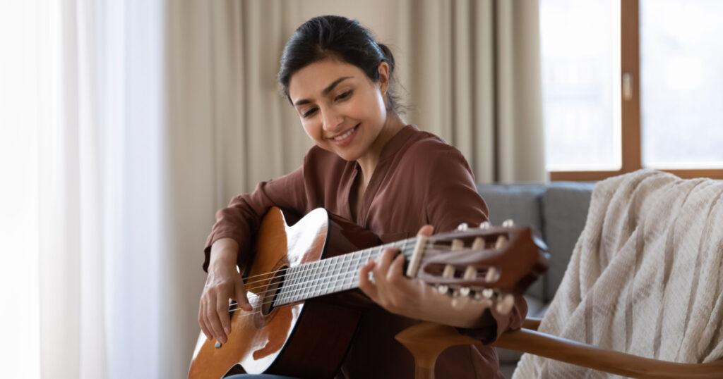 Creative hobby. Talented young mixed race female musician sit in armchair alone compose instrumental song using classic guitar. Smiling biracial lady play calm melody on musical instrument. Copy space
