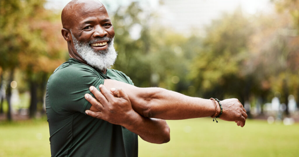 Senior fitness, exercise and black man stretching outdoor at park for energy, health and wellness in retirement. Portrait, face and smile of male in nature for workout, cardio and training in summer

