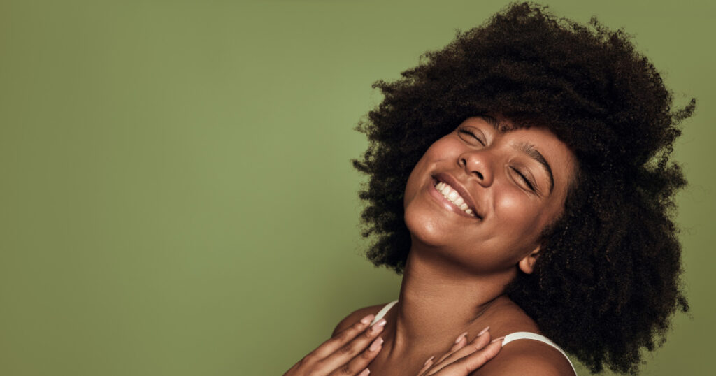 Side view of cheerful young African American female millennial with Coily hair and perfect skin smiling brightly with closed eyes against green background
