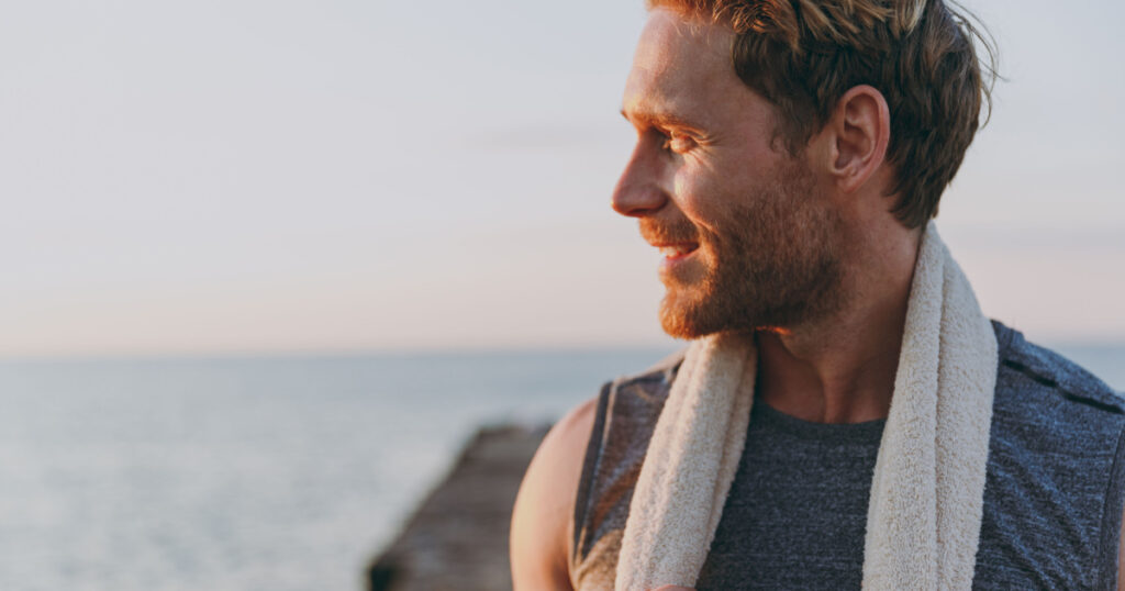 Close up happy young strong sporty athletic toned fit sportsman man in sports clothes towel warm up train look aside at sunrise sun dawn over sea beach outdoor on pier seaside in summer day morning.
