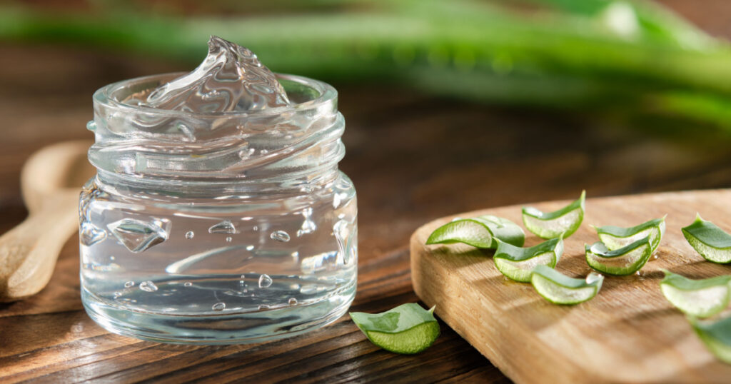 Transparent glass jar of aloe vera gel, sliced aloe leaves on a cutting board. Aloe vera plant on background. Natural cosmetic and medicinal remedy.
