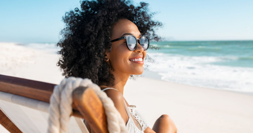 Happy young black woman relaxing on deck chair at beach wearing spectacles. Smiling african american girl with sunglasses enjoy vacation. Carefree happy young woman sunbathing at sea with copy space.
