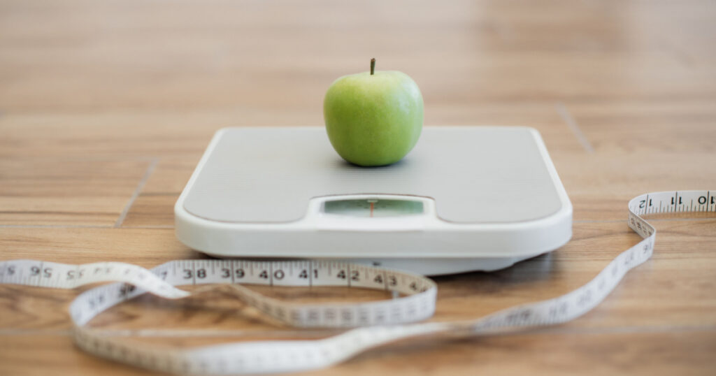 Close up view of green ripe apple being placed on bathroom scales while measuring tape lying on wooden floor nearby. Keeping active and eating balanced diet helping in maintaining healthy weight.
