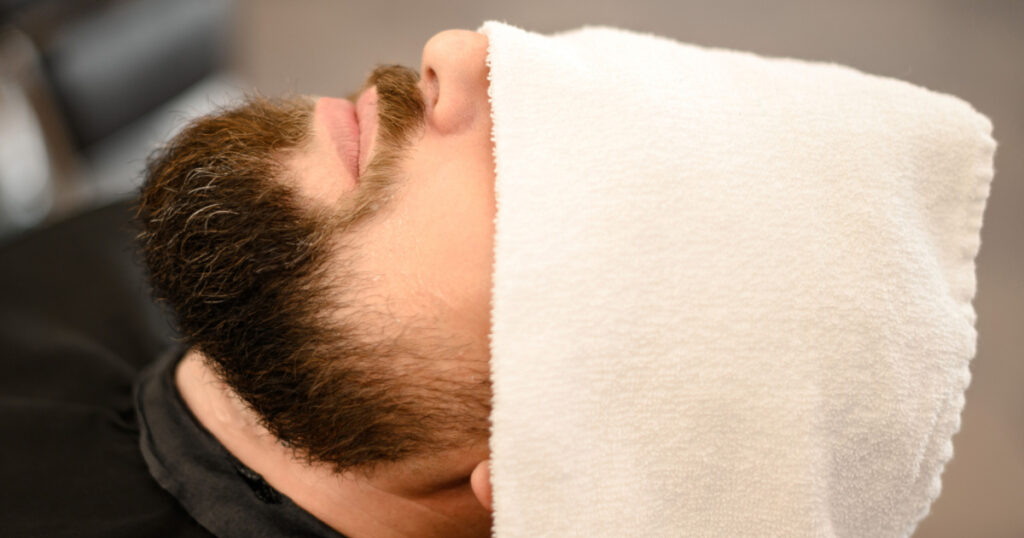A client is lying on a chair in a barbershop with his beard wrapped in a towel. A towel covers the mans eyes.
