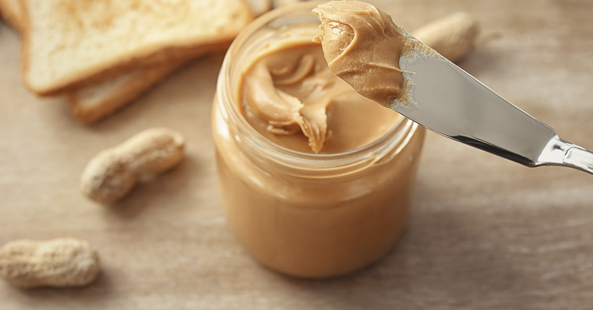 Knife being used in jar of peanut butter. Toast and whole peanuts on table beside jar of peanut butter.