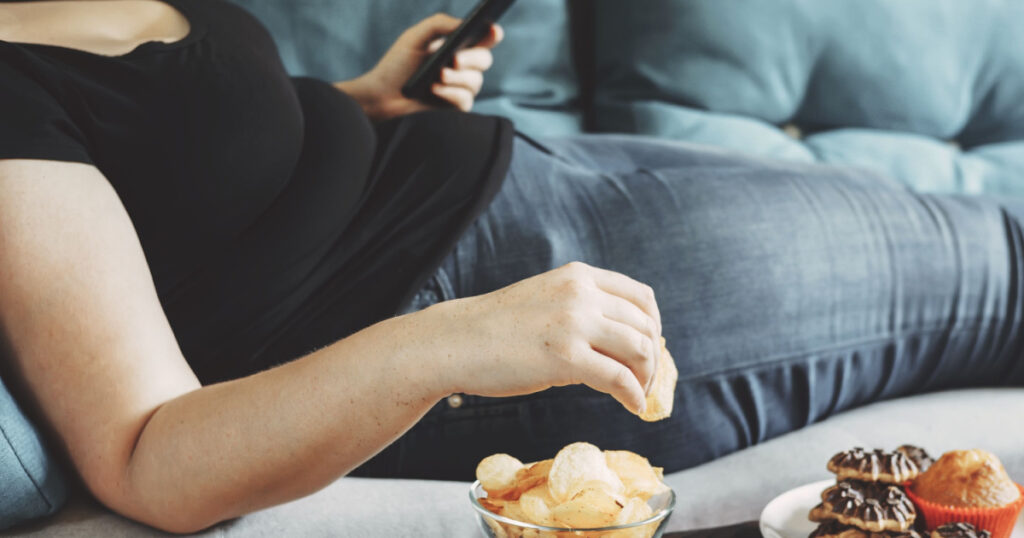g, sedentary lifestyle, compulsive overeating. Obese woman laying on sofa with smartphone eating chips
