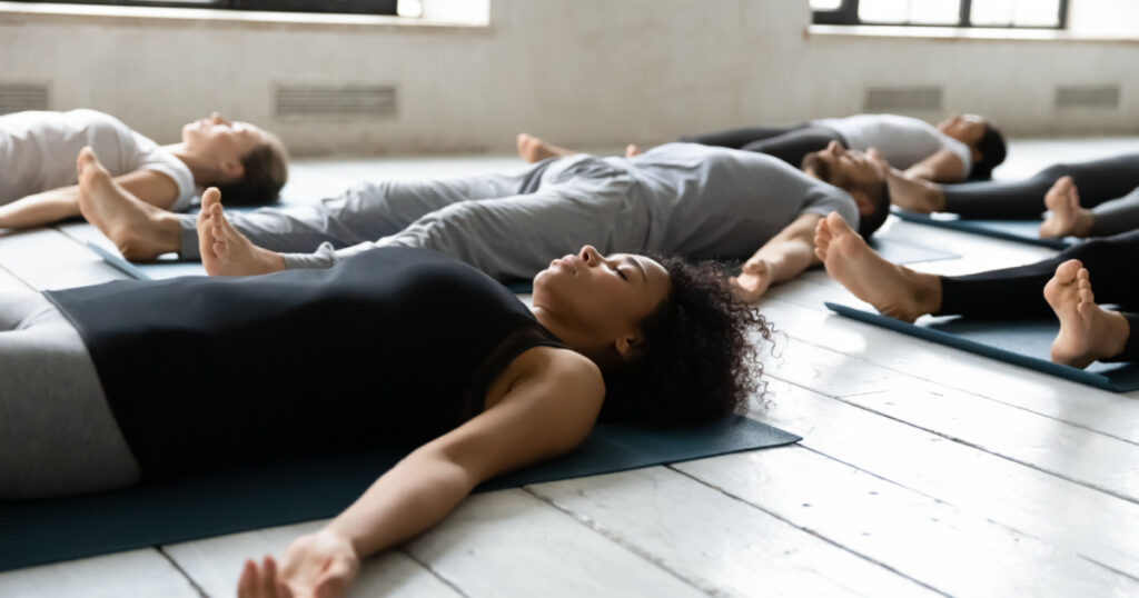 Diverse people resting after work out lying on carpets performing Shavasana or Corpse Dead position focus on African female, final pose of yoga class deep restoration, body and mind relaxation concept