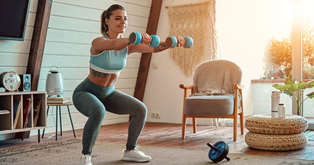 Beautiful young athletic girl in leggings and top crouches with dumbbells at home. Sport, healthy lifestyle.
